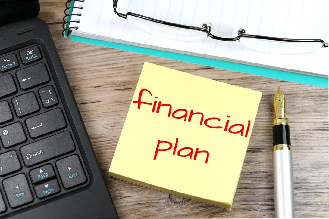 En gul kom-ihåg-lapp med texten 'Financial Plan' på ett skrivbord bredvid en laptop och en penna.