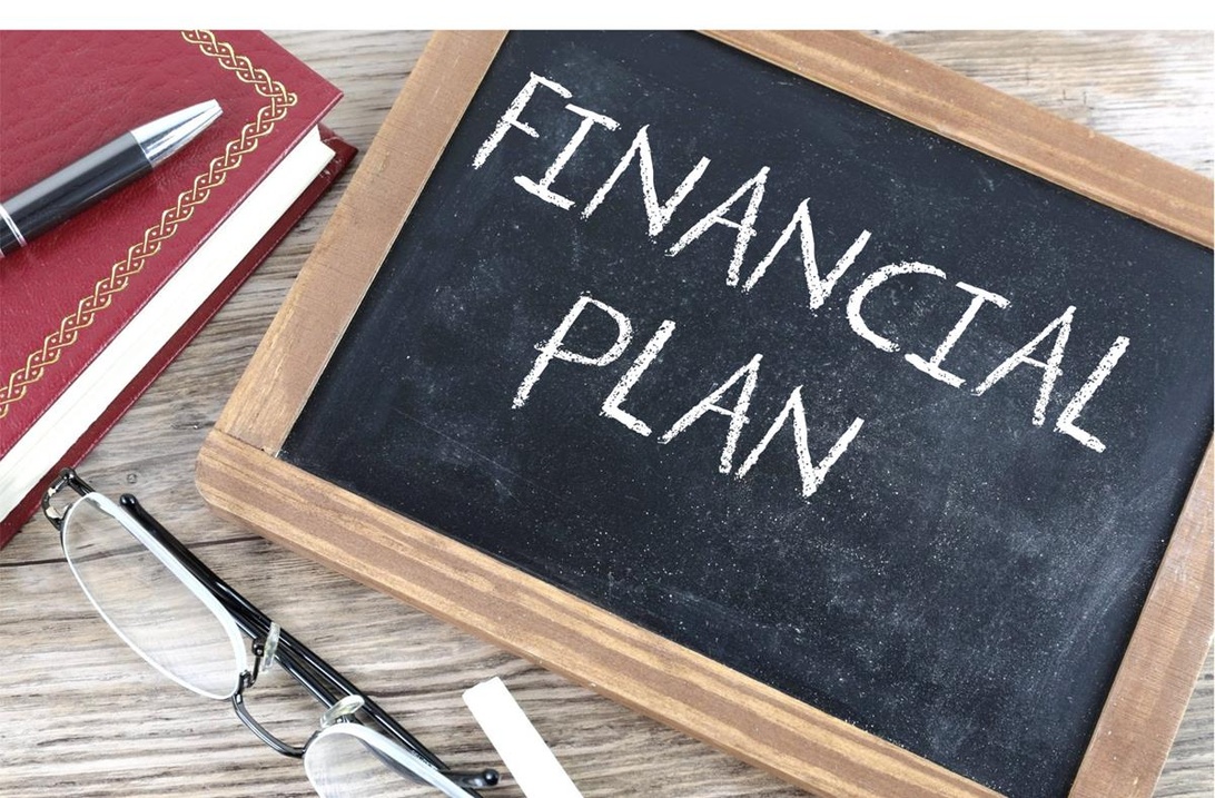 En svart tavla med texten 'FINANCIAL PLAN' skriven med krita, omgiven av anteckningsbok, penna och glasögon på ett träbord.